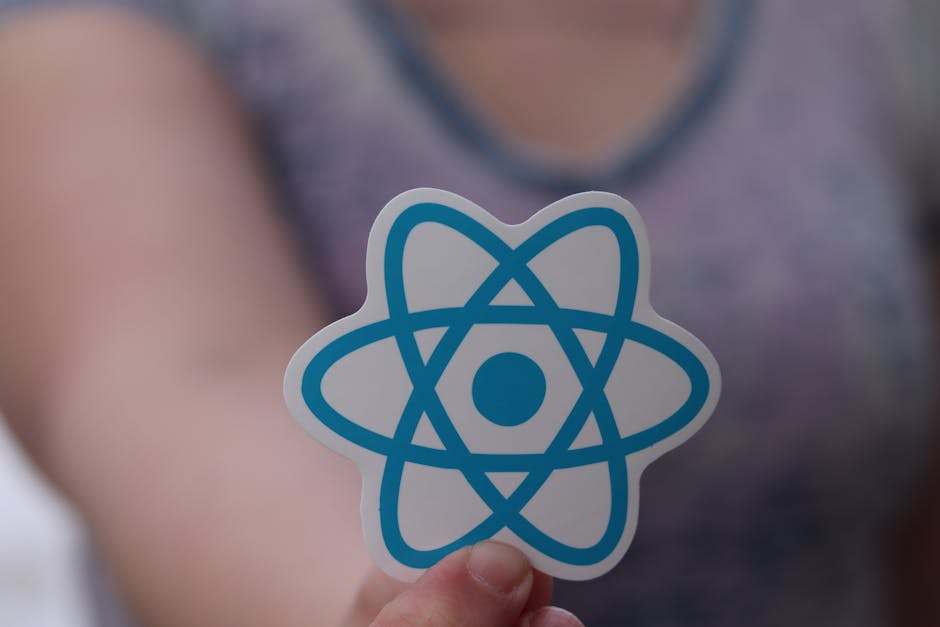 Close-up of a person's hand holding a white sticker with a blue atom symbol, set against a blurred background of a person wearing a grey tank top. The image is captured indoors with soft lighting, foc