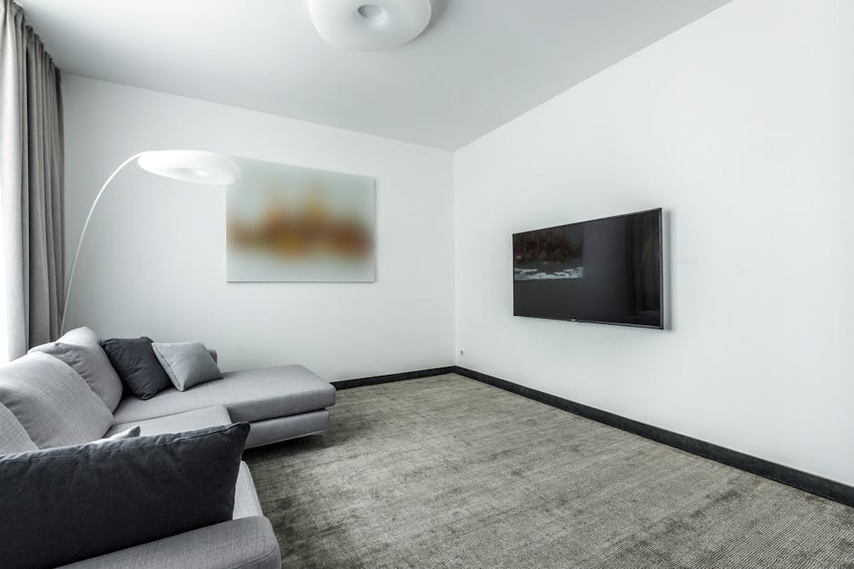 A yellow canister vacuum cleaner with a black hose and crevice attachment resting on an ornate, multicolored area rug in a living room. To the left, a wooden TV stand with black metal legs holds a fla